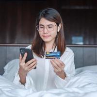 woman using mobile smart phone and credit card for online shopping while making order on bed in morning at home. technology, ecommerce, digital banking online payment and apartment living concept photo