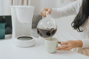 joven mujer feliz usando cafetera en casa. mujer asiática sirviendo café en una taza y haciendo café en casa. foto