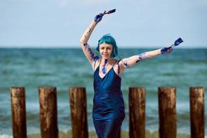 Artistic blue-haired woman performance artist smeared with blue gouache paints dancing on beach photo