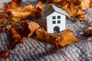 Autumnal Background. Toy house and dried orange fall maple leaves on grey knitted sweater. Thanksgiving banner copy space. Hygge mood cold weather concept. Hello Autumn with family photo