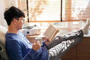 Handsome Asian Man wearing eyeglasses and smiling while Reading Book At Home living room lounge, literature and leisure concept photo