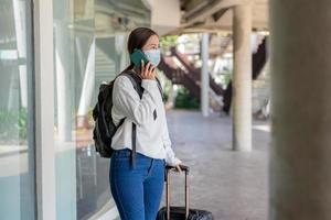 viaje, mujer con camisa blanca de manga larga y jeans, con una máscara. uso de teléfono móvil durante el viaje con maleta en la mano izquierda y mochila foto