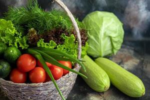 Fresh vegetables for salad in a basket. Tomatoes and cucumbers with zucchini and cabbage with dill. Spring harvest, benefits and vitamins. On a dark background. photo
