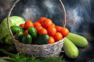 Fresh vegetables for salad in a basket. Tomatoes and cucumbers with zucchini and cabbage with dill. Spring harvest, benefits and vitamins. On a dark background. photo