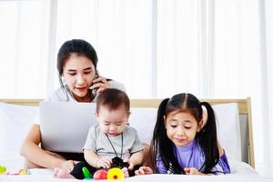 Baby and little daughter play toy on bed while Busy freelancer mother working on laptop photo