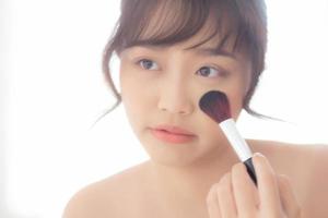 retrato de belleza joven mujer asiática sonríe con cara mirando espejo aplicando maquillaje con cepillo de mejilla en el dormitorio, hermosa chica sosteniendo colorete, cuidado de la piel y concepto cosmético. foto