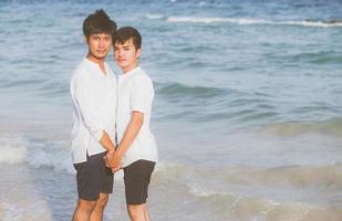 Homosexual portrait young asian couple standing together on beach in summer, asia gay holding hands going sea for leisure and relax with romantic and happy in vacation at sea, LGBT with legal concept. photo