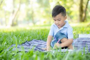 niño pequeño asiático leyendo y escribiendo un libro en el parque, tarea de niños de asia y estudio en el verano, el niño se relaja dibujando en el cuaderno en el concepto de vacaciones, educación y desarrollo. foto