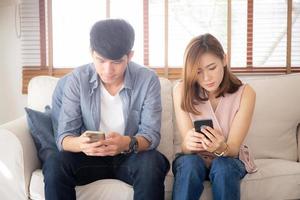 Young asian couple sitting on sofa with problem about relationship because addicts social network media together, asia family expression with looking phone with ignoring and indifferent. photo