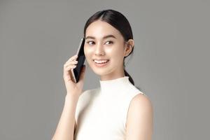 mujer asiática feliz sosteniendo el teléfono y hablando aislado sobre fondo gris. retrato de una chica hermosa en el estudio. foto