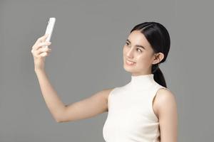 mujer asiática confiada sosteniendo teléfono y selfie aislado sobre fondo gris. retrato de una chica hermosa en el estudio. foto