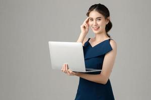 Young energetic Asian business woman holding laptop and thinking on gray background. Portrait of pretty girl in studio. Small business SME, freelance online, e-commerce Concept. photo