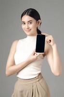 Confident Asian woman holding phone showing blank screen and looking at camera isolated on gray background. Portrait of a beautiful girl in the studio. photo