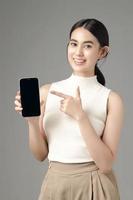 Confident Asian woman holding phone showing blank screen and looking at camera isolated on gray background. Portrait of a beautiful girl in the studio. photo