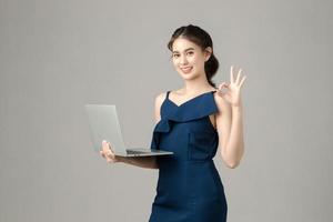 joven mujer de negocios asiática enérgica sosteniendo una computadora portátil que muestra una señal de ok sobre fondo gris. retrato de niña bonita en estudio. pyme de pequeñas empresas, autónomo en línea, concepto de comercio electrónico. foto