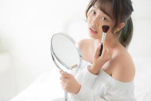 Beauty portrait young asian woman smile with face looking mirror applying makeup with brush cheek in the bedroom, beautiful of girl holding blusher, skin care and cosmetic concept. photo