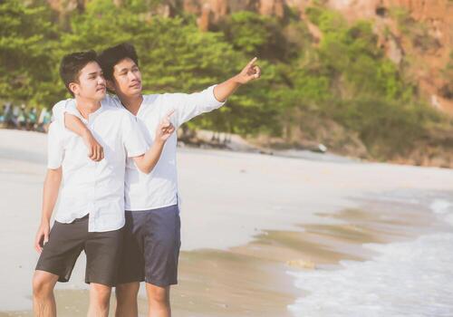 Homosexual portrait young asian couple standing pointing something together on beach in summer, asia gay going sea for leisure with romantic and happy in vacation at sea, LGBT with legal concept. photo