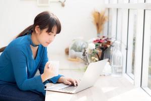 Beautiful young freelance asian woman smiling working and on laptop computer at desk coffee shop with professional, asia girl using notebook and drink coffee, business and lifestyle concept. photo