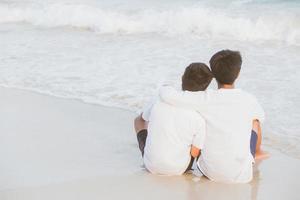 Back view homosexual portrait young asian couple sitting hug together on beach in summer, asia gay going tourism for leisure and relax with romantic and happy in vacation at sea, LGBT legal concept. photo