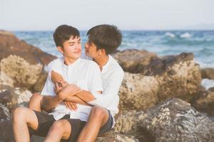 retrato homosexual joven pareja asiática sentada abrazada en roca o piedra en la playa en verano, turismo gay de asia para el ocio y relajarse con felicidad en vacaciones en el mar, concepto legal lgbt. foto