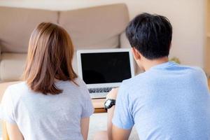 Back view of young asian couple working laptop with blank screen display, family planning and searching content together, man and woman looking computer, business and communication concept. photo