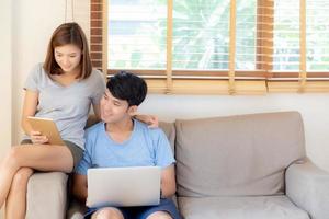 hermosa joven pareja asiática alegre independiente trabajando con un hombre usando una computadora portátil y una mujer usando una tableta en el sofá, la familia se relaja y comparte la comunicación juntos en casa, el concepto de estilo de vida. foto