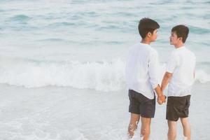 Back view young asian couple standing together on beach in summer, asia gay holding hands going sea for leisure and relax with romantic and happy in vacation at sea, LGBT with legal concept. photo