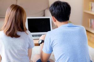 Back view of young asian couple working laptop with blank screen display, family planning and searching content together, man and woman looking computer, business and communication concept. photo