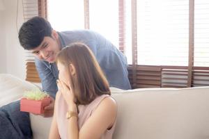 Young asian couple celebrate birthday together, asia man giving gift box present to woman for surprise at living room, female feeling happy and excited anniversary, holiday valentine concept. photo