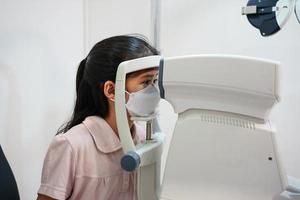 Ophthalmologist examining the eyes of an Asian girl in a clinic. They wear protective face masks. photo