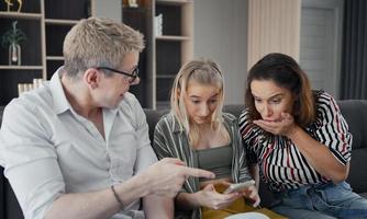 padres caucásicos sentados en el sofá y una hija emocionada usando un teléfono inteligente, están felices y preguntándose algo en la pantalla con una cara extraña en la sala de estar. foto