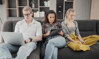 adicción a los gadgets. joven caucásico de tres sosteniendo y usando diferentes dispositivos electrónicos mientras está sentado en el sofá en la sala de estar en casa. padres y su hija con aparatos modernos foto