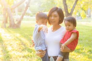 Beautiful young asian mother carrying little boy and girl in the park, asia woman happy having son and daughter and hug kid, mom love and embracing child together, mother day and family concept.