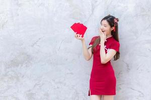 Asian beautiful woman in red dress stands and holds a red envelope with an excited expression on the grey background in Chinese new year theme. photo