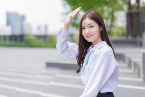 hermosa estudiante asiática de secundaria en el uniforme escolar con aparatos ortopédicos en los dientes, levantó la mano para bloquear la luz del sol y sonríe confiadamente feliz con la ciudad en el fondo. foto