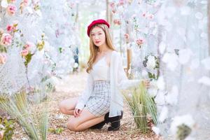 Asian woman has long bronze hair and wears red cap, white dress and black boots while she sits among flower garden in winter season. photo