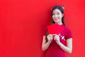 hermosa mujer asiática usa un cheongsam rojo y sostiene sobres rojos mientras mira a la cámara y sonríe alegremente con el fondo rojo. foto