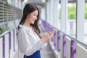 Professional beautiful confident young Asian businesswoman stands on flyover of skytrain outdoors in town while uses her smartphone to send messages to office.