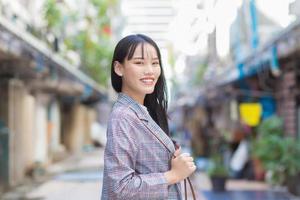 Confident young Asian woman who wears a Brown striped blazer and shoulder bag smiles happily and looks at the camera as she commute to work through the old town. photo