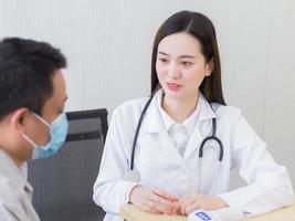 Professional beautiful young Asian female doctor talking with a man patient about his pain and symptom while they put on a face mask to prevent Coronavirus disease and Thermometer on table.3 photo