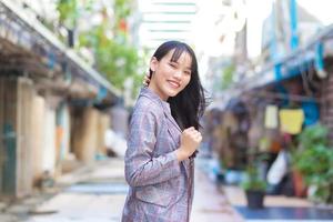 Confident young Asian female who wears a grey striped blazer and shoulder bag smiles happily and looks at the camera as she commute to work through the old town. photo