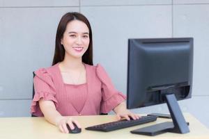 Professional young Asian business woman who wears pink dress is working while she sits  and smiles in an office typing on a keyboard and mouse. photo