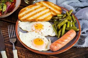 Breakfast with eggs, grilled sausage, green beans and toast on wooden background photo