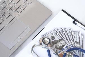 High angle view of stethoscope and dollar banknotes on clipboard with laptop keyboard on white marble tabletop, medical expense and insurance concept photo