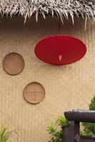 paraguas de papel rojo con 2 cestas de trilla en la pared de tejido de bambú de la cabaña tradicional con decoración de plantas verdes borrosas en primer plano en estilo vintage y marco vertical