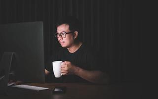 Early working in the morning concept, Asian man working on computer in the dark room and holding white coffee cup photo