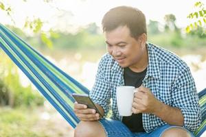 hombre sentado en una hamaca y sosteniendo una taza de café y un teléfono inteligente. tiro al aire libre con efecto de luz solar matutina foto