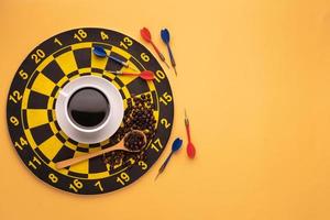 Cup of coffee on dart board on yellow background. Composition with free space for text or design photo