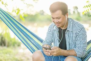 hombre sentado en una hamaca y usando un teléfono inteligente con auriculares blancos. tiro al aire libre con efecto de luz solar matutina foto