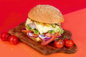 Delicious burger on the board with cutlet, tomatoes and cheese, cucumber and lettuce. On a red background. Fast food. photo